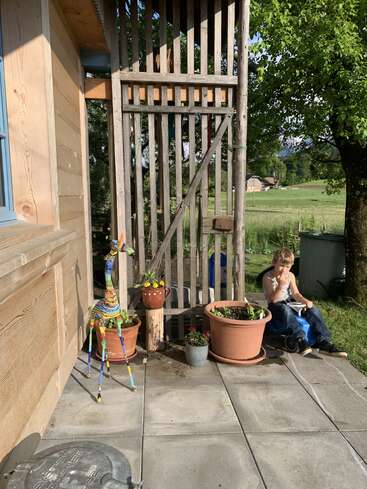 Un chico joven se sienta a comer en una silla azul al aire libre, junto a plantas en macetas, una estructura de madera, una colorida escultura de una jirafa, hierba verde y árboles bajo la luz del sol.