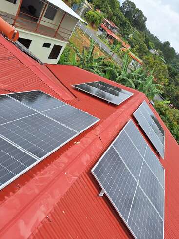 Hay seis paneles solares instalados en un tejado metálico rojo. Al fondo, hay árboles, vegetación, casas cercanas y una tranquila carretera en la ladera de una colina.