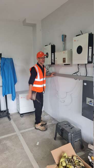 Un hombre con chaleco de seguridad naranja y gorra se encuentra en un cuarto de servicio, inspeccionando el equipo eléctrico montado en la pared. Cerca se ven herramientas y suministros.