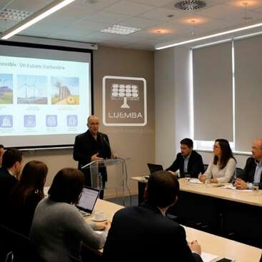 A man presents at a conference room, discussing renewable energy. Attendees listen attentively, some taking notes. A projector displays sustainability images and the LUEMBA logo.