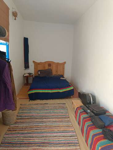 Ein einfaches, gemütliches Schlafzimmer mit einem Holzbett, einer bunt gestreiften Decke, einem Teppich und einer Bank. Minimale Dekoration, weiße Wände und ein kleines Fenster schaffen eine friedliche Atmosphäre.