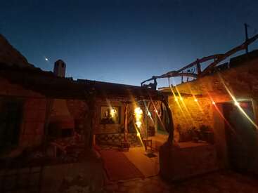 Ein gemütliches, rustikales Steinhaus leuchtet in der Abenddämmerung warm unter Lichterketten. Der tiefblaue Himmel kontrastiert mit dem einladenden, sanft beleuchteten Innenhof und den einfachen Möbeln.