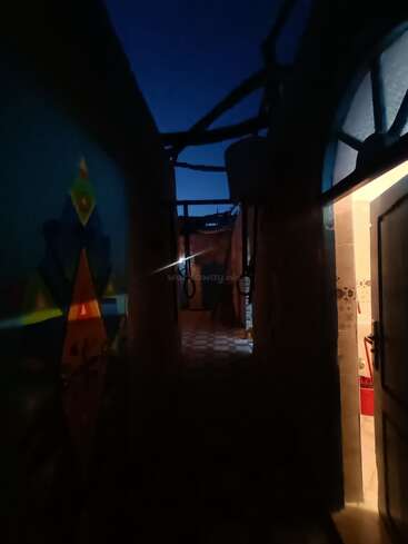 Ein schwach beleuchteter Korridor bei Nacht, mit geometrischer Wandkunst auf der linken Seite. Eine offene Tür gibt den Blick auf einen warmen, beleuchteten Raum auf der rechten Seite frei. Mysteriöse Atmosphäre.