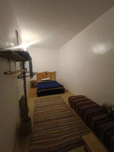 Dieses Bild zeigt ein kleines, einfaches Zimmer mit einem Einzelbett, einem Kopfteil aus Holz, einem gestreiften Teppich, einer Bank, einem Hängeregal und schlichten weißen Wänden, die sanft beleuchtet sind.