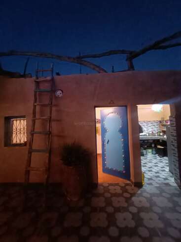 Uma casa rústica ao anoitecer apresenta uma porta azul decorativa, escada de madeira, vaso de plantas e azulejos estampados. Uma luz quente brilha em seu interior, criando um ambiente aconchegante.