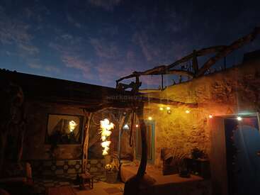 Une cour confortable au crépuscule, éclairée par des guirlandes lumineuses et des lanternes douces. Les murs en pierre rustique, les poutres en bois et le ciel paisible du soir créent une atmosphère magique.