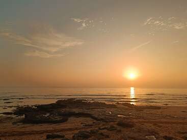 L'image sereine capture une plage calme au coucher du soleil, avec de douces vagues, des rochers éparpillés sur le sable, la lumière dorée du soleil et des nuages doux qui brillent dans le ciel chaud.