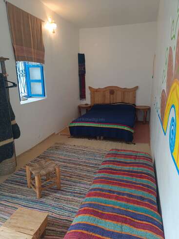 Une chambre étroite et confortable avec un lit simple, des couvertures rayées colorées, un banc en bois, un tapis tissé, un petit tabouret et une fenêtre avec des bordures bleues. Décor vivant.