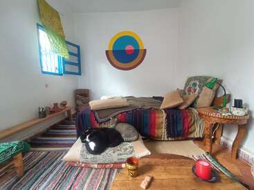 Ein gemütliches, farbenfrohes Zimmer mit einem Bett, gestreiften Decken, Kissen, einem Wandbild, einem offenen Fenster, einer auf Kissen schlafenden Katze, Kerzen, Teppichen und Holzmöbeln.