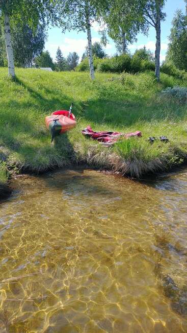 Un luminoso día de verano junto a un lago cristalino, con un kayak rojo, un remo, unos zapatos y una manta a rayas sobre la hierba verde, bajo unos abedules. Una escena relajante.
