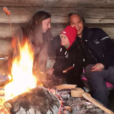Eine Familie sitzt gemeinsam an einem gemütlichen Lagerfeuer in einer rustikalen Blockhütte und lächelt einander warm an. Sie braten etwas am Feuer und genießen gemeinsam einen friedlichen, glücklichen Moment.