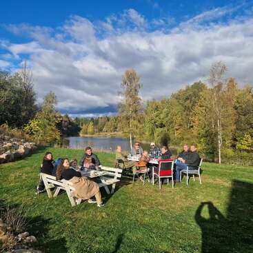 Eine Gruppe von Menschen sitzt im Freien auf Bänken an einem See und genießt den sonnigen Tag, umgeben von grünen Bäumen und einem malerischen, teilweise bewölkten Himmel.