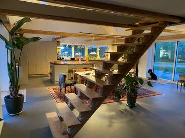 Moderner offener Küchen- und Essbereich mit Holztreppe, großen Fenstern, Zimmerpflanzen, sichtbaren Balken, gemütlicher Beleuchtung, Perserteppich und einem Hund an der Glasschiebetür.