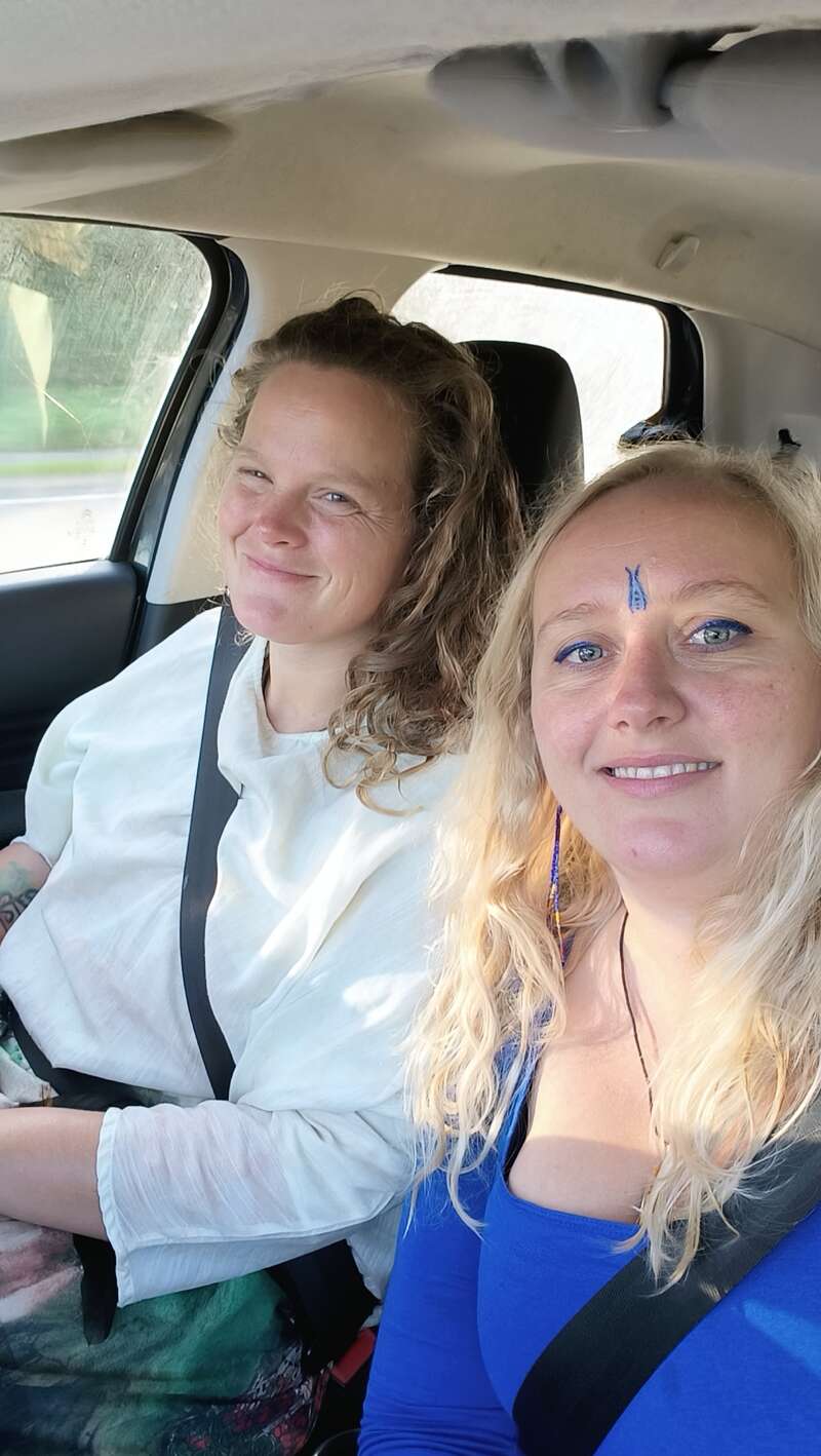 Deux femmes souriantes sont assises dans une voiture. L'une a les cheveux bouclés et porte sa ceinture de sécurité. L'autre a de longs cheveux blonds et du maquillage bleu sur le front.