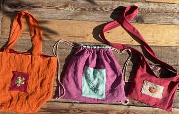 Three handmade fabric bags are displayed on a wooden surface: an orange tote, a pink drawstring backpack, and a red shoulder bag, each with decorated patches.