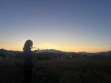Die Silhouette einer Person steht in der Abenddämmerung auf einem Feld und blickt auf die ruhigen Berge und die verblassenden Lichter der Stadt unter einem friedlichen Abendhimmel.