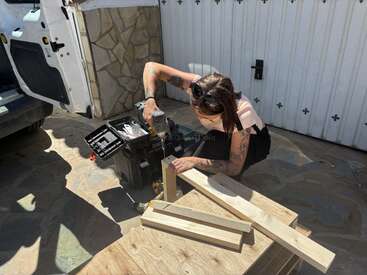 Eine Frau mit Tattoos montiert draußen mit einer Bohrmaschine Holzbretter. Sie arbeitet neben einem Werkzeugkasten in der Nähe eines weißen Garagentors und eines Lieferwagens.
