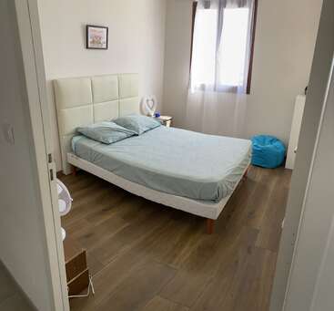 Un dormitorio ordenado con una cama azul claro, dos almohadas, mesilla de noche, puf azul, ventana grande, suelo de madera, decoración minimalista y arte en la pared.