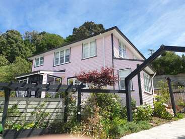 A imagem mostra uma casa de dois andares com exterior rosa, acabamento branco e telhado preto, cercada por um jardim exuberante e um céu azul.