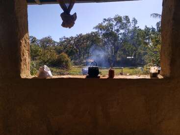 A rustic kitchen window overlooks a sunlit garden with trees, smoke rising from a kettle, a cup, and containers on the ledge, creating a peaceful scene.