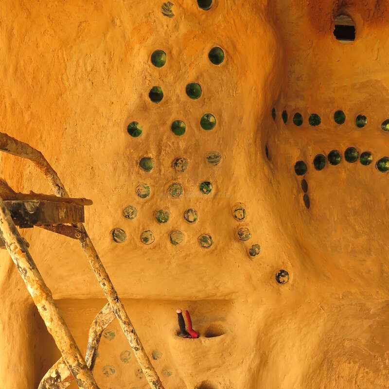 This image shows an earthen interior wall embedded with green glass bottles, a construction ladder covered in mud, and some tools, all bathed in warm sunlight.