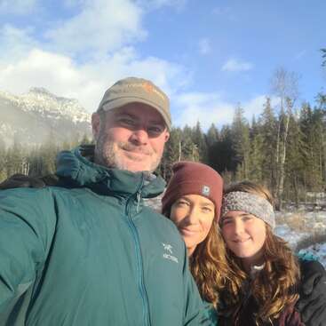 Uma família de três pessoas, bem agasalhada, posa ao ar livre em uma paisagem montanhosa ensolarada e coberta de neve, cercada por árvores perenes e um céu azul, curtindo uma aventura de inverno juntos.