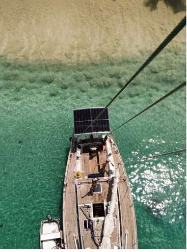 A sailboat is anchored in crystal-clear turquoise water near a sandy beach. The aerial view highlights the boat’s deck, solar panel, and inviting tropical scenery.