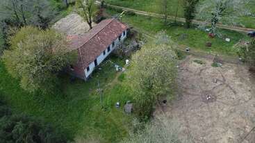 An aerial view shows a rustic house with a red-tiled roof, surrounded by green grass, trees, a dirt yard, and scattered outdoor objects, blending with nature.