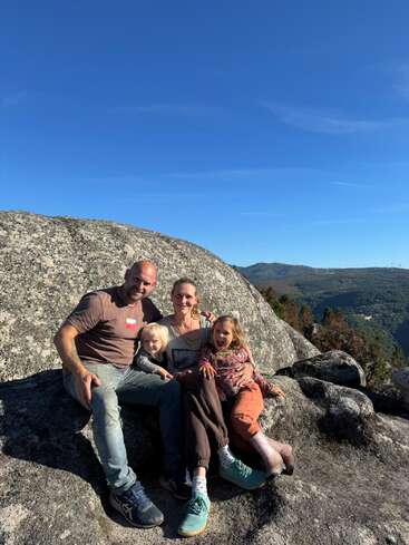 A happy family of four sits on large rocks under a clear blue sky, enjoying a scenic mountain view with greenery and distant hills in the background.