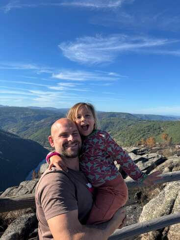 A man joyfully holds a young child outdoors. Both are smiling with mountains, forests, and a bright blue sky in the scenic background, radiating happiness.