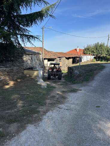 A rustic rural scene shows a small tractor parked by old stone buildings with red tile roofs, surrounded by greenery and a quiet, sunlit road.