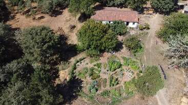 This aerial image shows a rustic house with a red-tiled roof surrounded by trees. In front, there’s a circular, well-maintained vegetable garden with pathways.