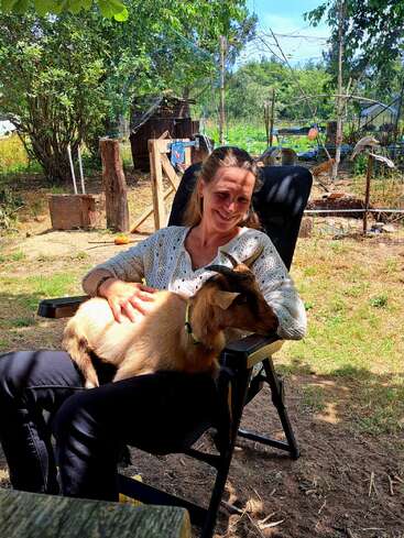 A woman sits outdoors in a black chair, smiling while holding a brown goat on her lap. The background includes trees, plants, and a rustic garden.