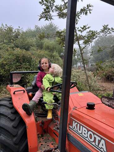 Two young children sit on an orange Kubota tractor outdoors. One child wears earmuffs, the other is dressed in green, both enjoying a misty, natural setting.