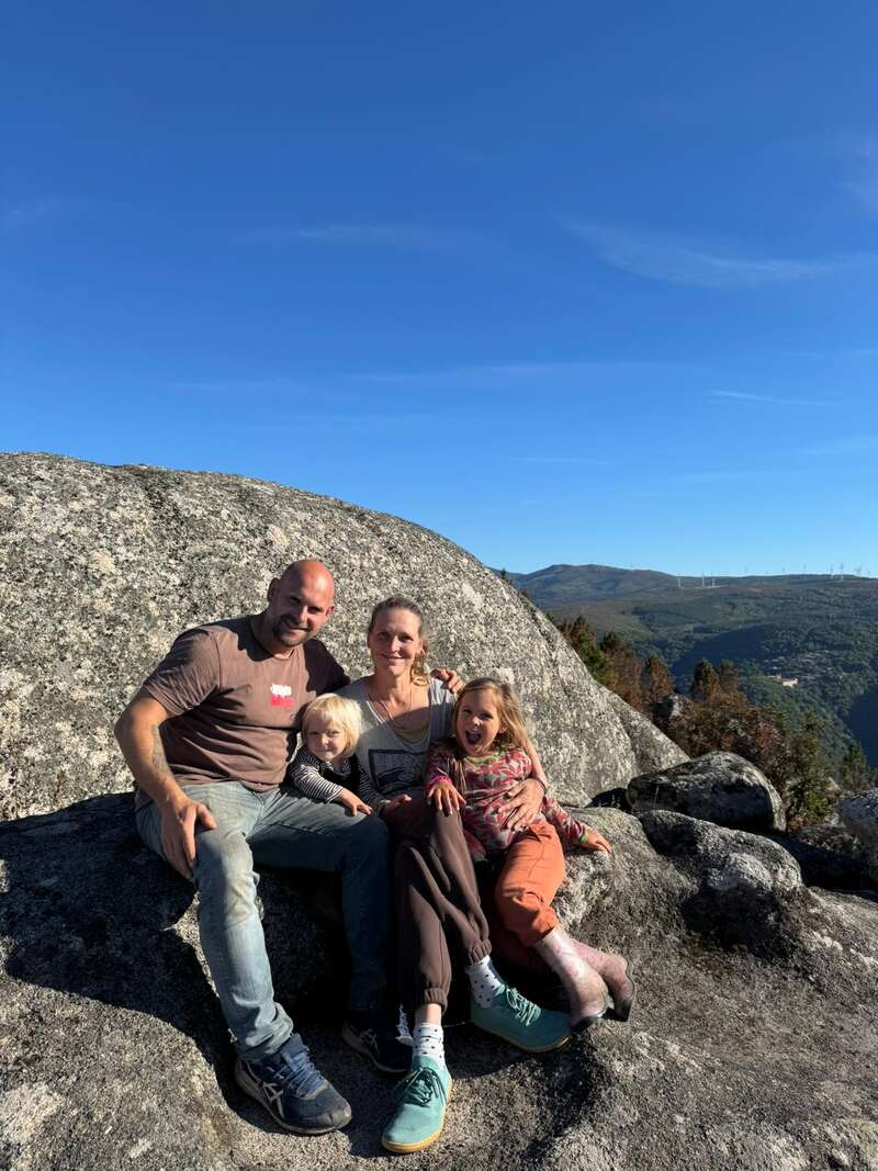 A happy family of four sits on large rocks under a clear blue sky, enjoying a scenic mountain view with greenery and distant hills in the background.