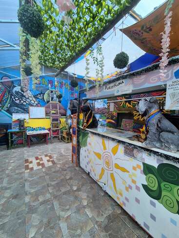 A vibrant, artistic indoor space with colorful murals, hanging plants, and creative decor. A counter labeled "PuriyKuna" features a dog statue and cheerful, sunny designs.