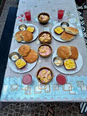 A neatly set breakfast table with buns, scrambled eggs, butter, banana and yogurt bowls, and glasses of red juice on a floral-patterned tablecloth.