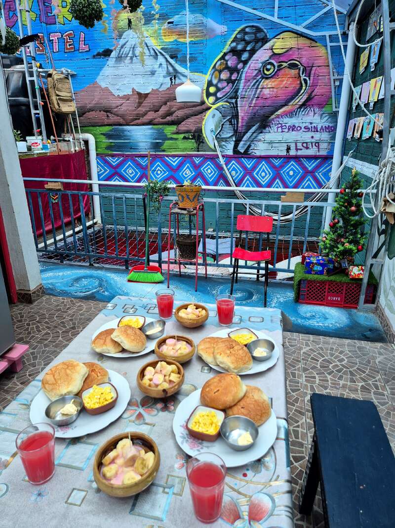 A colorful outdoor setting with a breakfast table features bread, scrambled eggs, fruit salad, and juice. Bright mural of a bird and mountain in background.