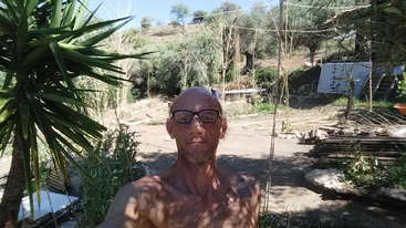 La imagen representa a un hombre de pie delante de un árbol, con una masa de agua detrás. Lleva sombrero y bigote, y el fondo es un entorno natural.