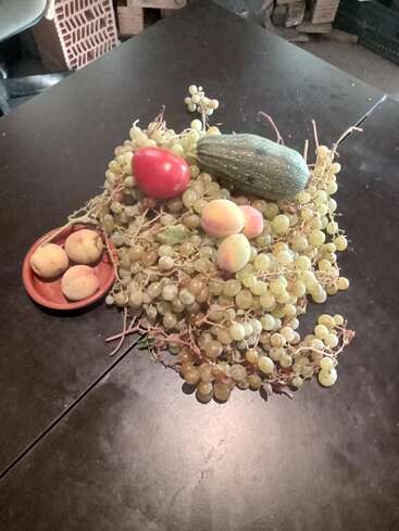 A still life arrangement with green grapes, peaches, a tomato, a zucchini, and three apricots on a small brown plate, all on a dark table.