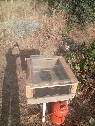 Una cámara casera de vidrio y madera se encuentra al aire libre sobre hierba seca, conectada a una bombona de gas naranja, posiblemente utilizada para calefacción o experimentos científicos. Se ve la sombra de una persona.
