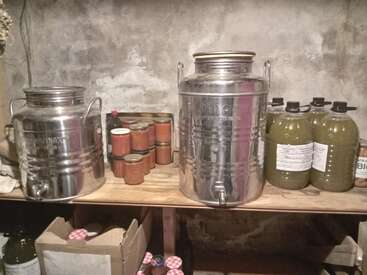 Esta imagen muestra una estantería rústica de despensa con grandes recipientes metálicos, tarros de cristal de conservas, botellas de plástico de líquido y una caja de cartón con tapas de tarros.