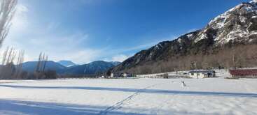 A imagem retrata uma paisagem serena de neve com uma cadeia de montanhas ao fundo, com uma pequena aldeia e um céu azul com nuvens finas.