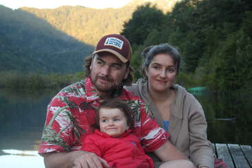 A imagem mostra uma família de três pessoas posando para uma foto, com um homem, uma mulher e uma criança, em um cenário de árvores e montanhas.