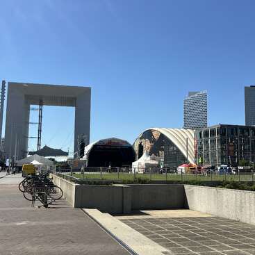 Paisagem urbana moderna com a Grande Arche, um palco ao ar livre, tendas e alguns arranha-céus ao fundo. Há bicicletas estacionadas ao longo de uma calçada ensolarada.