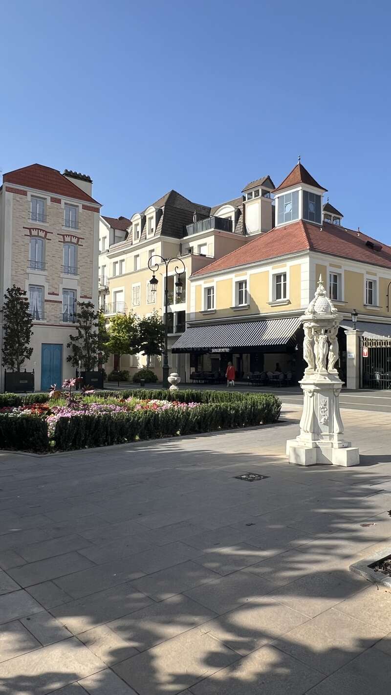 Uma pitoresca cena de rua europeia mostra prédios coloridos, uma estátua branca decorativa, jardins bem cuidados com flores, um céu azul claro e pessoas aproveitando um dia ensolarado.