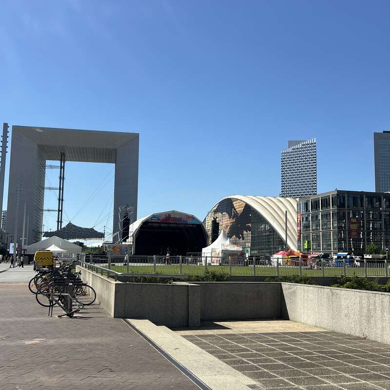 Paisagem urbana moderna com a Grande Arche, um palco ao ar livre, tendas e alguns arranha-céus ao fundo. Há bicicletas estacionadas ao longo de uma calçada ensolarada.