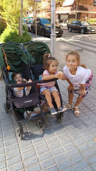 The image depicts two children in a stroller on a sidewalk, with a girl standing beside it, holding an ice cream cone, in a sunny urban setting.