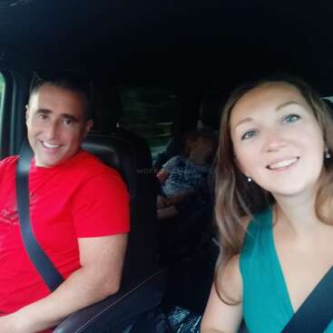 The image shows a man and woman in the front seats of a car, with a third person in the back seat, all wearing seatbelts and smiling at the camera.