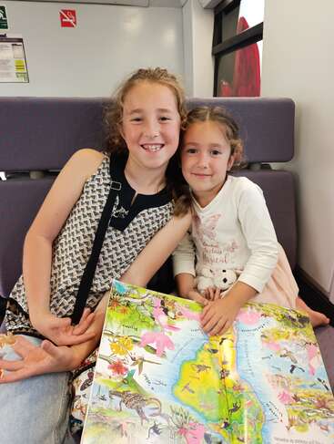 Two smiling young girls sit together on a train seat, holding a colorful illustrated book open. The atmosphere is cheerful, cozy, and filled with curiosity and joy.